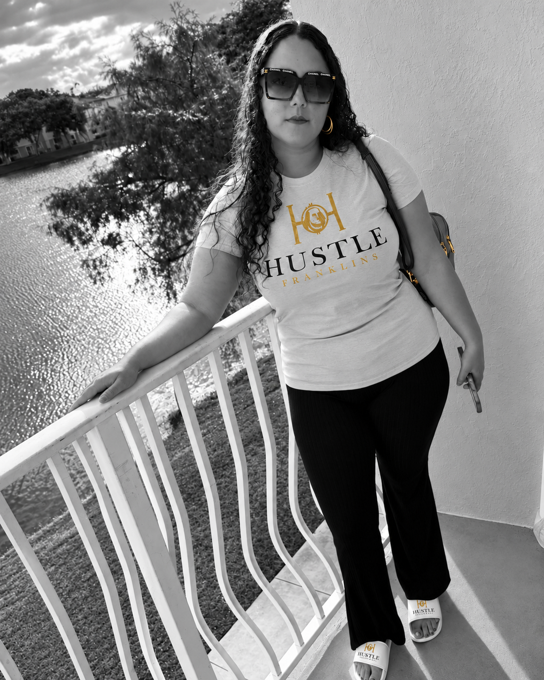 Woman wearing a Hustle Franklins tee on a balcony in a Noir and Gold campaign edit.