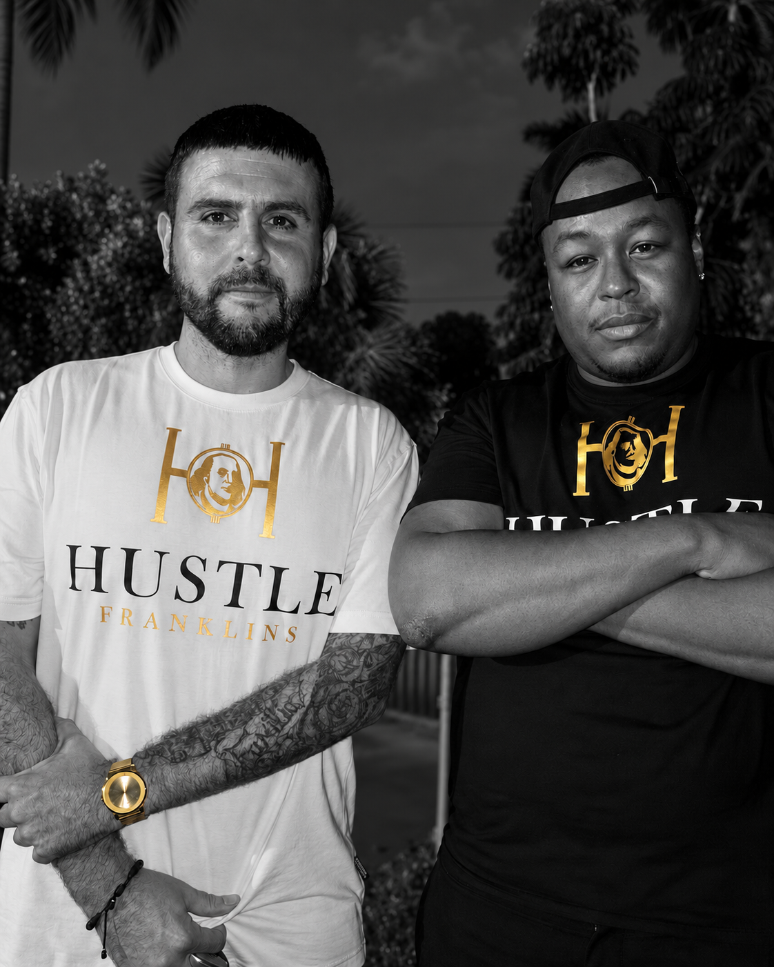 Two people wearing black and white Hustle Franklins tees with gold details at night.